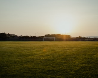 A tranquil football field at sunset, with a goalpost in the distance and lush green grass covering the ground. The sun is low on the horizon, casting a golden glow over the entire scene.
