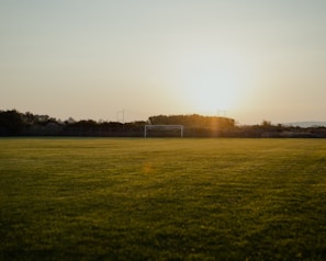 A tranquil football field at sunset, with a goalpost in the distance and lush green grass covering the ground. The sun is low on the horizon, casting a golden glow over the entire scene.