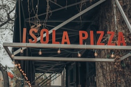A large sign with the words 'Isola Pizza' in bold, red letters is prominently displayed. It is mounted on a metal framework adorned with string lights. The background features a wall with creeping vines and a muted, natural aesthetic.