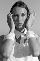 A black and white portrait of a person with sleek hair, wearing a button-up shirt with a pointed collar. Their hands are raised, lightly touching their face near the temples. The expression is serious and contemplative, creating a classic and timeless feel.