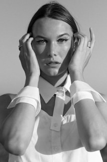 A black and white portrait of a person with sleek hair, wearing a button-up shirt with a pointed collar. Their hands are raised, lightly touching their face near the temples. The expression is serious and contemplative, creating a classic and timeless feel.