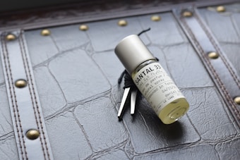 A bottle of Santal 33 perfume lies on a textured surface with a leather-like appearance. The bottle is cylindrical, clear, and has a minimalist label with black text. Metal elements and stitching adorn the surface, adding a vintage or classic aesthetic.