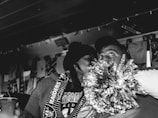 A black and white photograph captures a close moment between two people wearing fan gear. The person on the left is holding a pom-pom and appears to be engaged in an affectionate gesture towards the other person, who is looking upward with an expressive gaze. The background is adorned with banners and items suggesting a sports event or fan gathering.