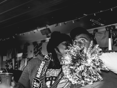 A black and white photograph captures a close moment between two people wearing fan gear. The person on the left is holding a pom-pom and appears to be engaged in an affectionate gesture towards the other person, who is looking upward with an expressive gaze. The background is adorned with banners and items suggesting a sports event or fan gathering.