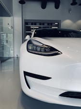 A sleek, modern electric car parked indoors, showcasing its aerodynamic design and clean lines. The image focuses on the left headlight and front portion of the vehicle, emphasizing its futuristic aesthetic.