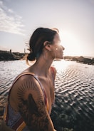 A woman with a large tattoo on her arm is sitting by a body of water during sunset. She has a serene expression and is wearing a multicolored striped swimsuit. Large hoop earrings accentuate her look.