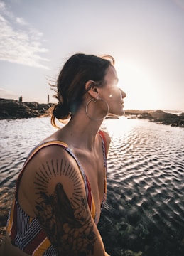 A woman with a large tattoo on her arm is sitting by a body of water during sunset. She has a serene expression and is wearing a multicolored striped swimsuit. Large hoop earrings accentuate her look.