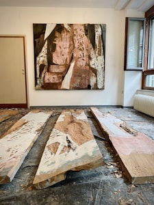The image depicts an art gallery room with a large abstract painting on the wall, featuring a mix of pink, brown, black, and white tones with intricate patterns and textures. On the floor, there are three long, wooden planks with painted designs that mirror the painting's aesthetic, showing layers of paint and carved details. The room is well-lit with natural light entering through a large open window.