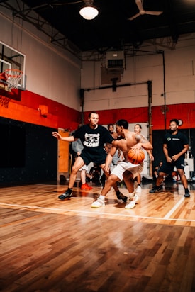 A group of men are playing basketball indoors on a polished wooden court. One player is dribbling the ball while others are actively engaged in the game, possibly trying to defend. The court is bordered by dark and red walls with a basketball hoop mounted on one side. The setting suggests an intense and energetic atmosphere.