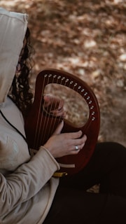 A person wearing a hooded jacket is playing a small, wooden harp with one hand. The background is blurred, showing an earthy, outdoor setting. The person is focused on the instrument, and their hair falls loosely around the hood.