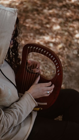A person wearing a hooded jacket is playing a small, wooden harp with one hand. The background is blurred, showing an earthy, outdoor setting. The person is focused on the instrument, and their hair falls loosely around the hood.