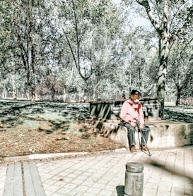 An older person wearing a pink sweater and cap sits on a concrete bench in a sunlit park. The surroundings include lush green trees, a paved walkway, and shadows cast by the sunlight filtering through the foliage.