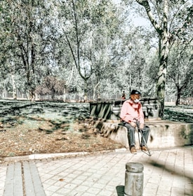 An older person wearing a pink sweater and cap sits on a concrete bench in a sunlit park. The surroundings include lush green trees, a paved walkway, and shadows cast by the sunlight filtering through the foliage.