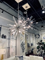 A stylish interior featuring two modern spiky chandeliers with bright lights hanging from a high ceiling. The room includes a white sofa with decorative cushions, a variety of wall art including circular and geometric designs, and several plants adding a touch of greenery. The space is well-lit, creating a sophisticated and contemporary atmosphere.
