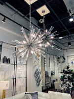 A stylish interior featuring two modern spiky chandeliers with bright lights hanging from a high ceiling. The room includes a white sofa with decorative cushions, a variety of wall art including circular and geometric designs, and several plants adding a touch of greenery. The space is well-lit, creating a sophisticated and contemporary atmosphere.