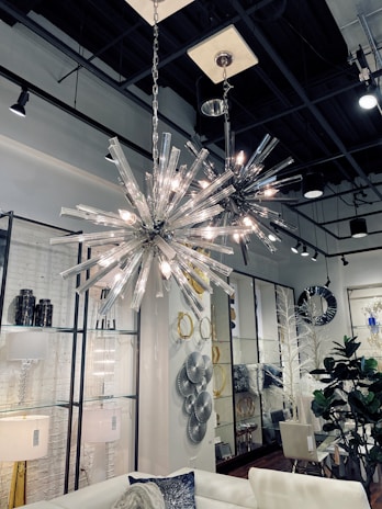 A stylish interior featuring two modern spiky chandeliers with bright lights hanging from a high ceiling. The room includes a white sofa with decorative cushions, a variety of wall art including circular and geometric designs, and several plants adding a touch of greenery. The space is well-lit, creating a sophisticated and contemporary atmosphere.