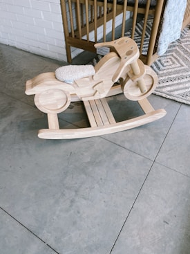 A wooden rocking motorcycle designed for children is placed on a tiled floor, adjacent to a wooden crib and a patterned carpet. The rocking toy features a light wooden finish and a small cushioned seat.