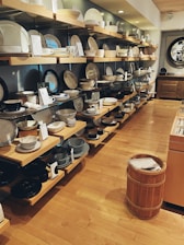 A well-organized retail display featuring various kitchenware items such as plates, bowls, and cups. The shelves are neatly arranged along a wall, showcasing different sizes and styles of dishware. The setting has a warm, inviting atmosphere with wooden floors and soft lighting.