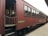 A vintage passenger train car painted in red with wooden paneling and several windows. The train is stationary on tracks next to a platform. The side of the car has a sign that reads 'Blue Bird'. The setting suggests an old-fashioned or historical railway experience.