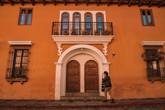 A person walks in front of a vibrant orange building with colonial architecture, featuring an arched wooden door, wrought iron balconies with potted flowers, and two upper windows on either side. The person is holding a guitar and is dressed casually.