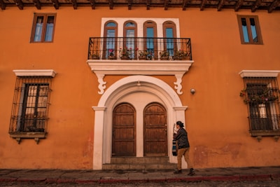A person walks in front of a vibrant orange building with colonial architecture, featuring an arched wooden door, wrought iron balconies with potted flowers, and two upper windows on either side. The person is holding a guitar and is dressed casually.