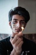 A young person is holding a lit match in the foreground, with words 'I AM FIRE' written on their face using adhesive tape and matchsticks. The focus is on the match, and the background is softly blurred.