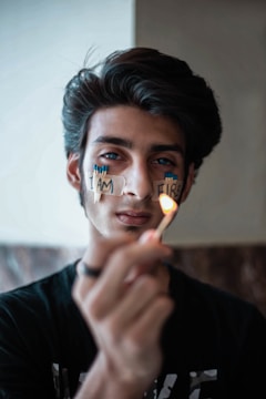 A young person is holding a lit match in the foreground, with words 'I AM FIRE' written on their face using adhesive tape and matchsticks. The focus is on the match, and the background is softly blurred.