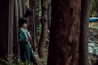 A person in a green, elaborate costume with a high top hat leans against a wooden structure in a forested area. The individual is holding a small book and appears to be deep in thought. Tall trees surround them, casting shadows and creating a secluded atmosphere.