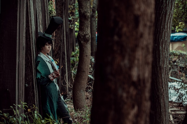 A person in a green, elaborate costume with a high top hat leans against a wooden structure in a forested area. The individual is holding a small book and appears to be deep in thought. Tall trees surround them, casting shadows and creating a secluded atmosphere.