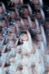 A visually complex image featuring a repeated pattern of a person in a white, sequined outfit. The person appears to be holding a reflective object, creating a kaleidoscope effect. The entire scene is adorned with sparkles and soft highlights, giving it a surreal and dreamy feel.