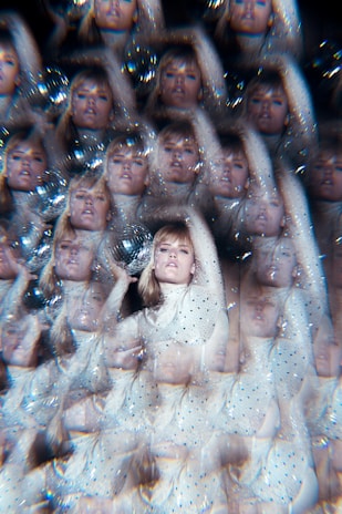 A visually complex image featuring a repeated pattern of a person in a white, sequined outfit. The person appears to be holding a reflective object, creating a kaleidoscope effect. The entire scene is adorned with sparkles and soft highlights, giving it a surreal and dreamy feel.