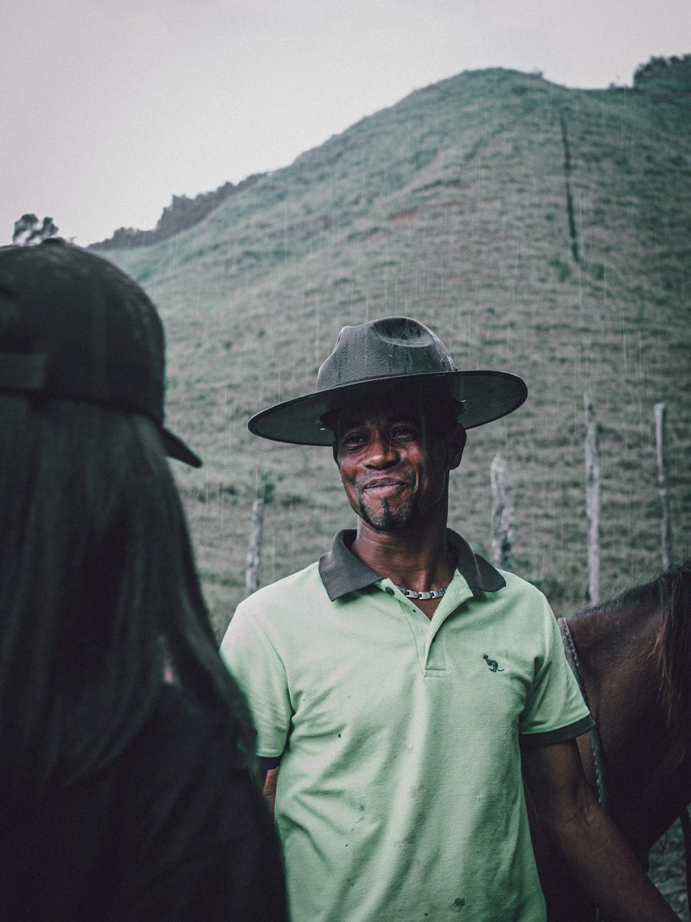 man in green polo shirt wearing black hat standing beside brown horse during daytime