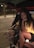A person with long curly hair is sitting in the back of a car with an open hatch. They are smiling and resting their chin on their hand. Next to them is a dog wearing a service dog vest. The setting appears to be a dimly lit parking lot at night.