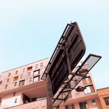 A large urban billboard stands in front of a modern multi-story building with numerous windows. The clear sky provides a stark contrast to the dark structure of the billboard, which is devoid of any advertising material.