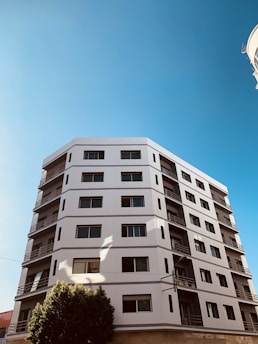 A modern, multi-story residential building with a geometric design, featuring a white facade and numerous windows. The structure is highlighted against a clear blue sky, with a tree partially visible in the foreground.