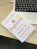 A book titled 'Sapiens: A Brief History of Humankind' lies on a wooden table next to the keyboard area of a laptop. The book has a clean white cover with red and black text, accompanied by a gray and gold seal. The laptop is open, showing a portion of the keyboard and the touchpad.