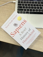 A book titled 'Sapiens: A Brief History of Humankind' lies on a wooden table next to the keyboard area of a laptop. The book has a clean white cover with red and black text, accompanied by a gray and gold seal. The laptop is open, showing a portion of the keyboard and the touchpad.