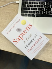 A book titled 'Sapiens: A Brief History of Humankind' lies on a wooden table next to the keyboard area of a laptop. The book has a clean white cover with red and black text, accompanied by a gray and gold seal. The laptop is open, showing a portion of the keyboard and the touchpad.