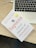 A book titled 'Sapiens: A Brief History of Humankind' lies on a wooden table next to the keyboard area of a laptop. The book has a clean white cover with red and black text, accompanied by a gray and gold seal. The laptop is open, showing a portion of the keyboard and the touchpad.
