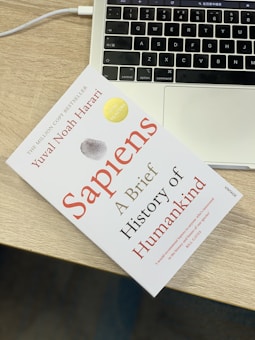 A book titled 'Sapiens: A Brief History of Humankind' lies on a wooden table next to the keyboard area of a laptop. The book has a clean white cover with red and black text, accompanied by a gray and gold seal. The laptop is open, showing a portion of the keyboard and the touchpad.