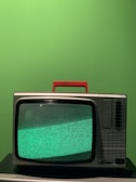 A vintage television set with a red handle sits on a black surface against a bright green background. The screen displays a static-filled, greenish noise pattern.