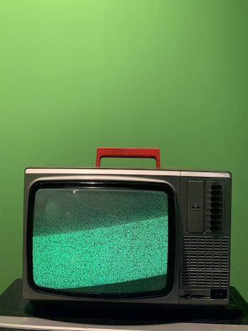 A vintage television set with a red handle sits on a black surface against a bright green background. The screen displays a static-filled, greenish noise pattern.