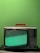 A vintage television set with a red handle sits on a black surface against a bright green background. The screen displays a static-filled, greenish noise pattern.