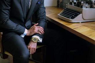 A person in a dark suit is seated at a wooden desk with an antique typewriter. The individual has accessorized with a gold watch and a ring, and their hands rest on their lap.