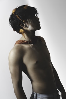 A shirtless person is posed in profile view with a focus on their muscular structure. Their head is slightly tilted back, and their hair is adorned with small feathers. The lighting creates dramatic shadows, accentuating the contours of their body.