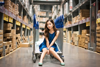 A young woman wearing a blue dress is sitting on a flatbed cart in a large warehouse aisle filled with stacked boxes. The floor is reflective, and the area is well-lit by overhead lighting. Blue handles of the carts can be seen behind her.