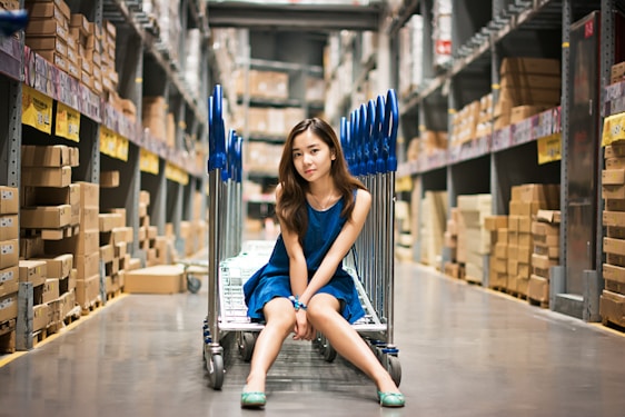A young woman wearing a blue dress is sitting on a flatbed cart in a large warehouse aisle filled with stacked boxes. The floor is reflective, and the area is well-lit by overhead lighting. Blue handles of the carts can be seen behind her.