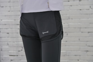 A person is wearing black athletic shorts over black leggings. The background features a white brick wall. Part of a white jacket with a zipper is visible at the top.