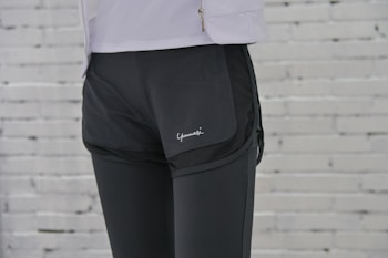 A person is wearing black athletic shorts over black leggings. The background features a white brick wall. Part of a white jacket with a zipper is visible at the top.