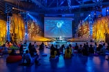 A large indoor space with people seated on the floor, facing a central stage where a performer is surrounded by colorful lighting. The room is decorated with large plants and nature-themed murals. Visuals on a big screen enhance the atmosphere.
